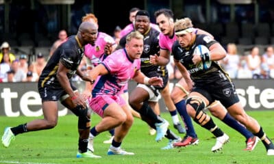 In the image, a dynamic moment from a rugby match is captured, showcasing a player in a vibrant pink kit, driving forward with the ball against the opposition. Surrounding him are players from the opposing team, dressed in black kits, who are intensely focused on halting his advance. The intensity and focus of the players are palpable, highlighting the physicality and fast-paced action typical of a high-stakes rugby game. This scene perfectly illustrates the competitive spirit and athleticism inherent in the sport.