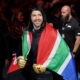 A South African MMA fighter draped in the national flag, wearing gold gloves, smiles as he makes his entrance at a UFC event, surrounded by his team and security.