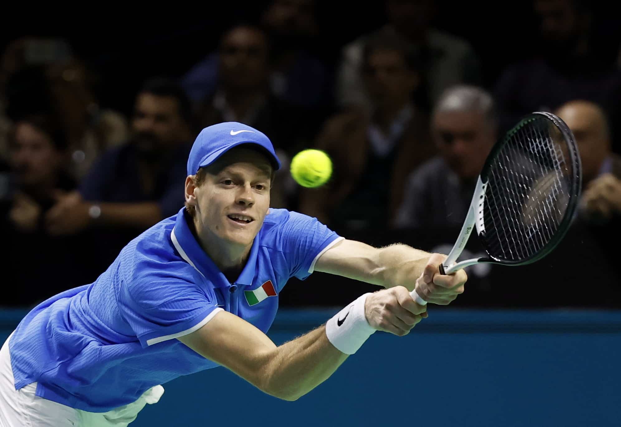 This image captures a tennis player in intense action during a match, dressed in a bright blue shirt with a national flag emblem, suggesting he's playing in a significant international event. He is focused on a low, forceful backhand stroke, showing excellent form and concentration. The blurred background emphasizes the speed and dynamism of the game, highlighting the player's athletic prowess and the high stakes of the competition.