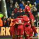 Members of Liverpool FC celebrate together on the pitch during a match, with one player lifted in the air amid the group. This joyful moment highlights team unity and excitement after a goal.