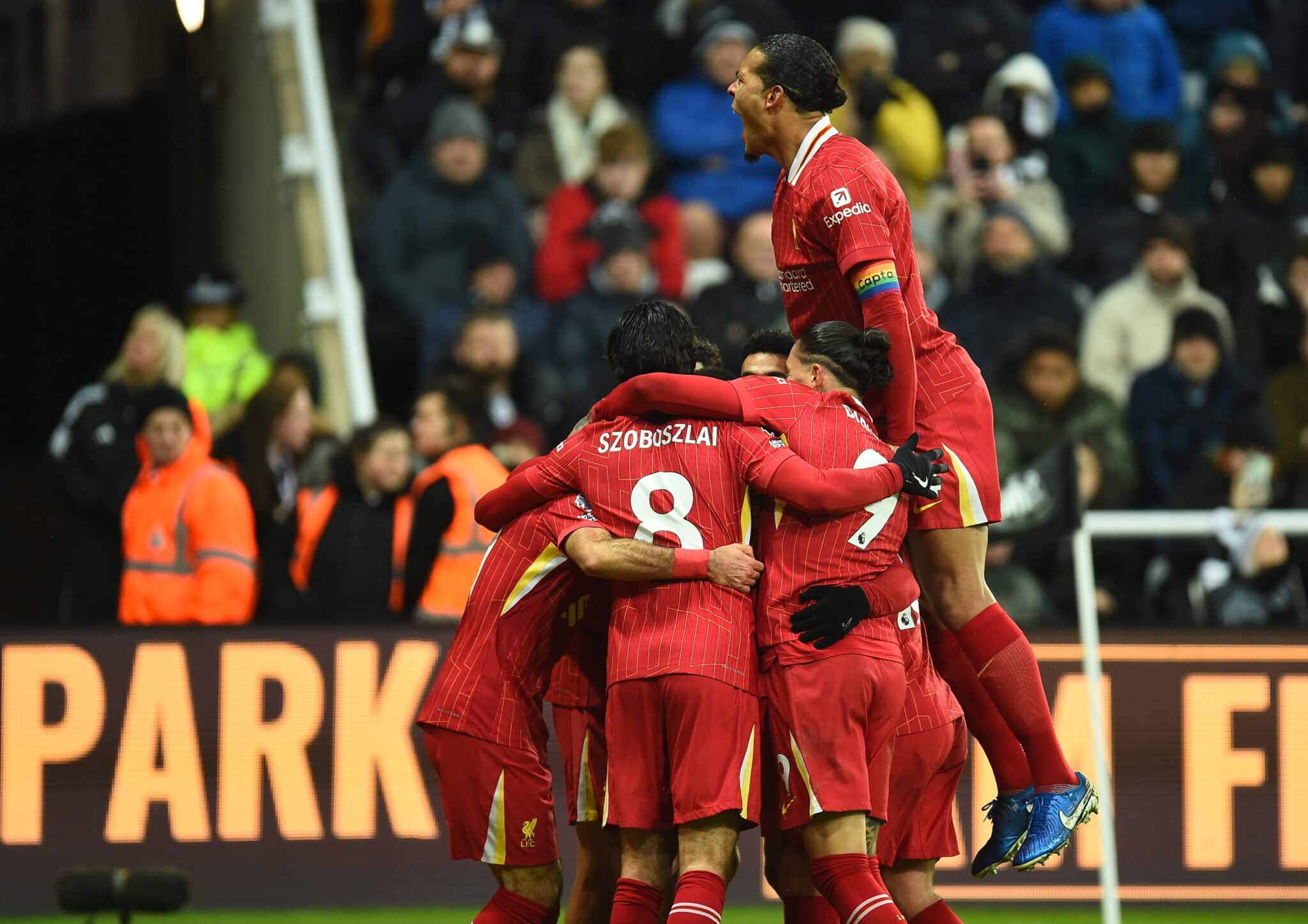 Members of Liverpool FC celebrate together on the pitch during a match, with one player lifted in the air amid the group. This joyful moment highlights team unity and excitement after a goal.