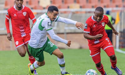 The image captures an intense moment from a football match, featuring three players in a tight contest for the ball. Two players, dressed in red uniforms, are from the same team, while the third player, in a green and white uniform, represents the opposition. The player in green is actively trying to maneuver around his opponents, displaying agility and focus. The backdrop shows a stadium, suggesting the scene is during a competitive league game. The athletes' expressions and body postures indicate the high stakes and fast-paced nature of the match.