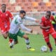 The image captures an intense moment from a football match, featuring three players in a tight contest for the ball. Two players, dressed in red uniforms, are from the same team, while the third player, in a green and white uniform, represents the opposition. The player in green is actively trying to maneuver around his opponents, displaying agility and focus. The backdrop shows a stadium, suggesting the scene is during a competitive league game. The athletes' expressions and body postures indicate the high stakes and fast-paced nature of the match.