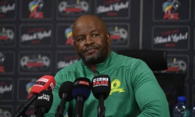 This image shows a man, presumably a coach, at a press conference wearing a Mamelodi Sundowns green top. Surrounded by microphones and logos from news outlets like SABC and EWN, he appears to be engaged in a significant media event related to the football club.