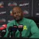 This image shows a man, presumably a coach, at a press conference wearing a Mamelodi Sundowns green top. Surrounded by microphones and logos from news outlets like SABC and EWN, he appears to be engaged in a significant media event related to the football club.