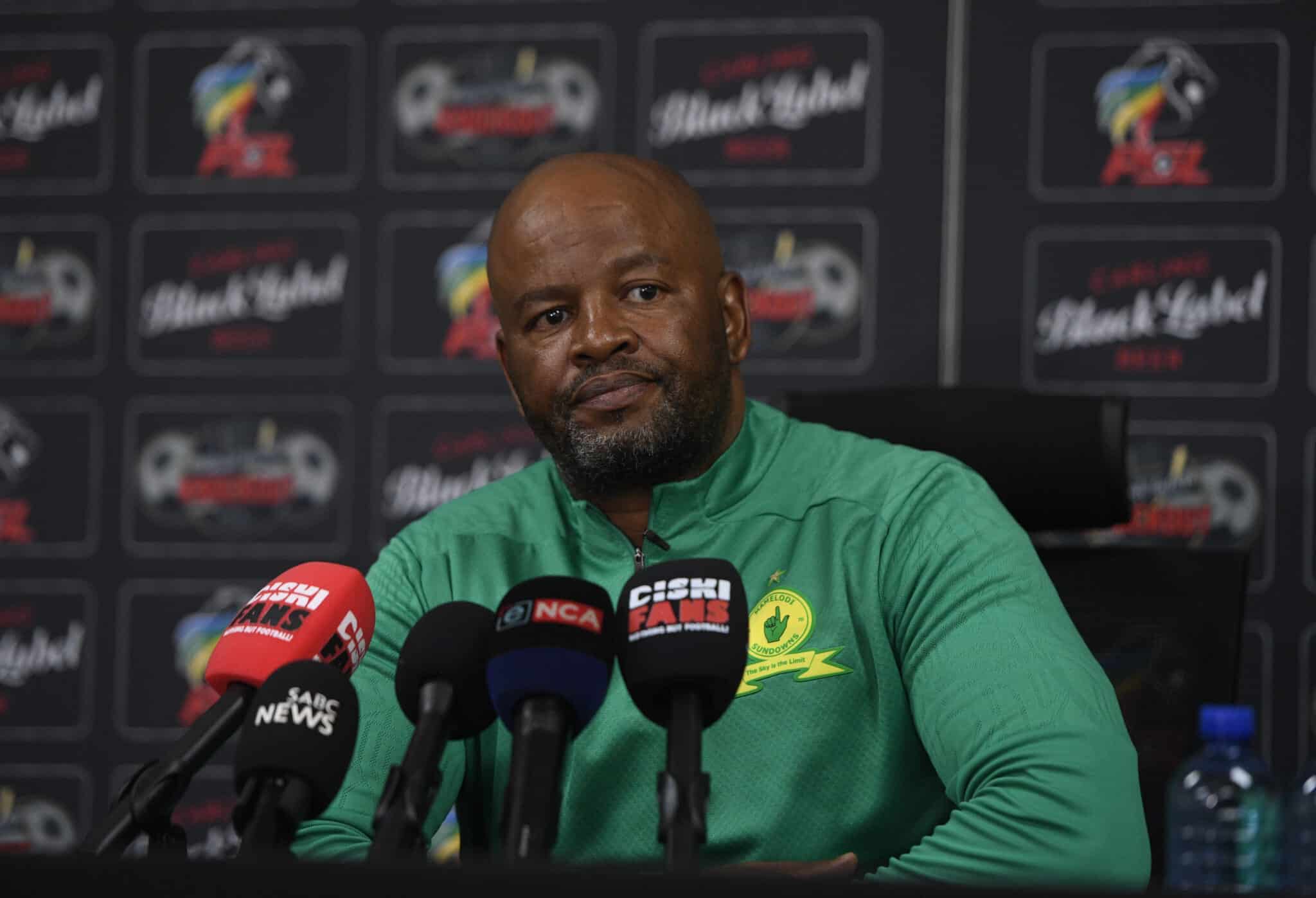 This image shows a man, presumably a coach, at a press conference wearing a Mamelodi Sundowns green top. Surrounded by microphones and logos from news outlets like SABC and EWN, he appears to be engaged in a significant media event related to the football club.