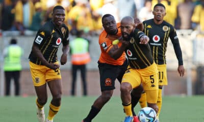 In the image, a moment from a soccer match between two teams, one in black and gold uniforms and the other in orange, captures a tense competition for the ball. The player in orange is tightly defending against a player from the opposing team, who is making a determined effort to maintain possession. This scene vividly illustrates the physical and strategic elements of soccer, showcasing the players' intense focus and athletic skill during a crucial match play.