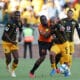 In the image, a moment from a soccer match between two teams, one in black and gold uniforms and the other in orange, captures a tense competition for the ball. The player in orange is tightly defending against a player from the opposing team, who is making a determined effort to maintain possession. This scene vividly illustrates the physical and strategic elements of soccer, showcasing the players' intense focus and athletic skill during a crucial match play.