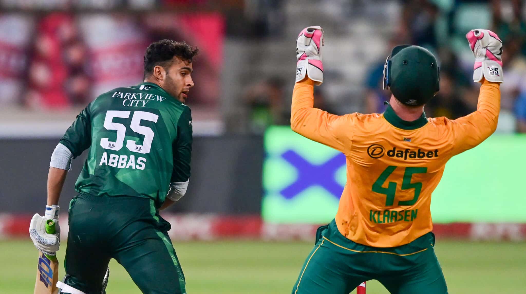 The image shows a moment from a cricket match with a batsman and a wicketkeeper in action. The batsman, dressed in a green jersey labeled "Abbas" and the number 55, is likely in the process of taking a run or reacting to a play. The wicketkeeper, in a vibrant orange kit with the number 45, is energetically raising his glove, possibly celebrating a dismissal or directing his teammates. The scene captures the intensity and excitement typical of a cricket game, highlighting player interactions and vibrant team colors.