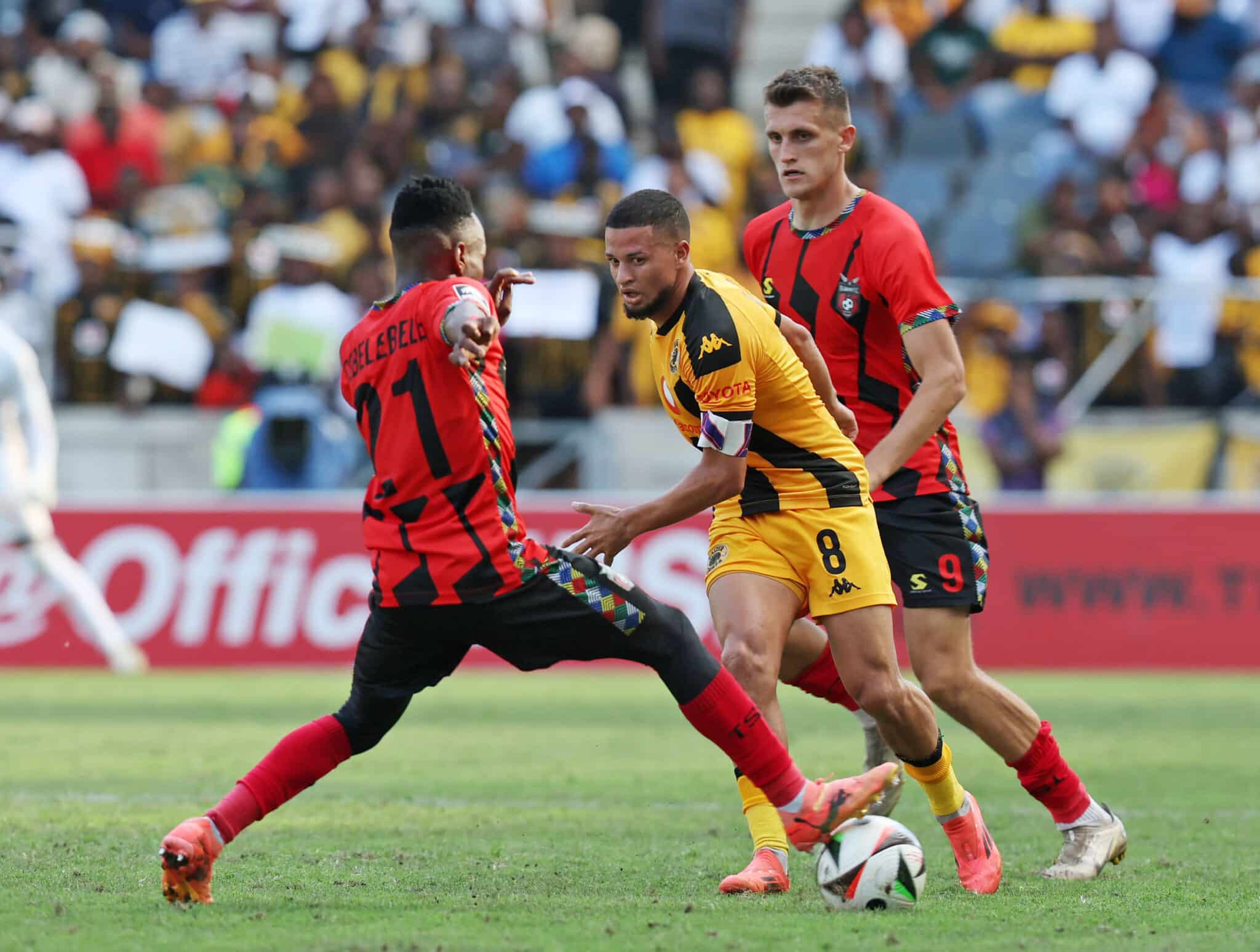 Dynamic action during a football match featuring Kaizer Chiefs and a rival team, with players intensely contesting the ball.