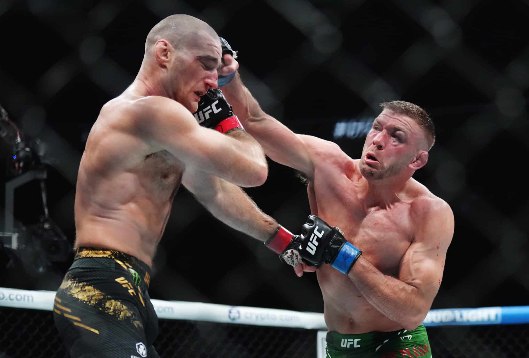 In the image, two UFC fighters are intensely engaged in combat during a match. One fighter, wearing black and gold shorts, is landing a powerful punch on his opponent, who is in green shorts. The impact of the punch is visibly jarring, demonstrating the high stakes and fierce competition in UFC fights. Both athletes display concentration and physical prowess, capturing the dramatic and brutal essence of mixed martial arts competitions.